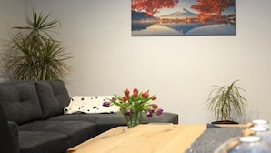 A living room with a dark grey sofa, a wooden coffee table, a vase with red and purple flowers, and a painting of a landscape with trees and a mountain. - Modern vacation apartment in a quiet location (Meißner)