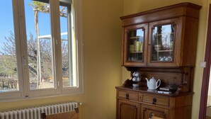 A round wooden dining table with a glass bowl centerpiece, surrounded by wicker chairs. A wooden cabinet with glass doors is to the right, and a window with a view of trees and a building is to the left. - Country house ! 5 minutes from the village center. (Caromb)