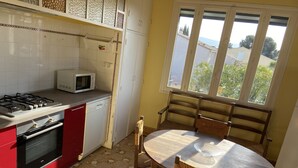 A kitchen with a gas stove, microwave, and dishwasher. There is a dining table with chairs and a window with a view of buildings and trees. - Country house ! 5 minutes from the village center. (Caromb)