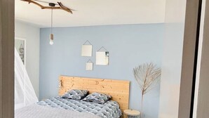 A bedroom with a wooden headboard, a bed with patterned bedding, a hanging light fixture, and a small side table with a plant. - Face a le Mer Fleury (Fleury)