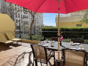 An outdoor patio with a table set for two, surrounded by wicker chairs and a red umbrella.