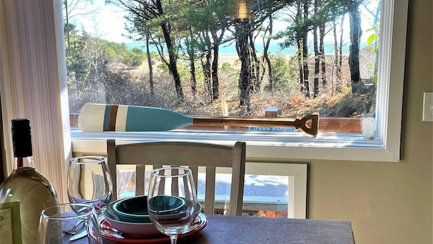 A dining table with wine glasses, plates, and a bottle, set against a window with a view of trees and a paddleboard. - 🐚Shell City on Lt Island (Wellfleet)