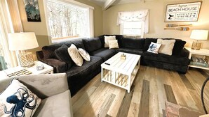 A cozy living room with a sectional sofa, a coffee table, and a large window offering a view of trees. - 🐚Shell City on Lt Island (Wellfleet)