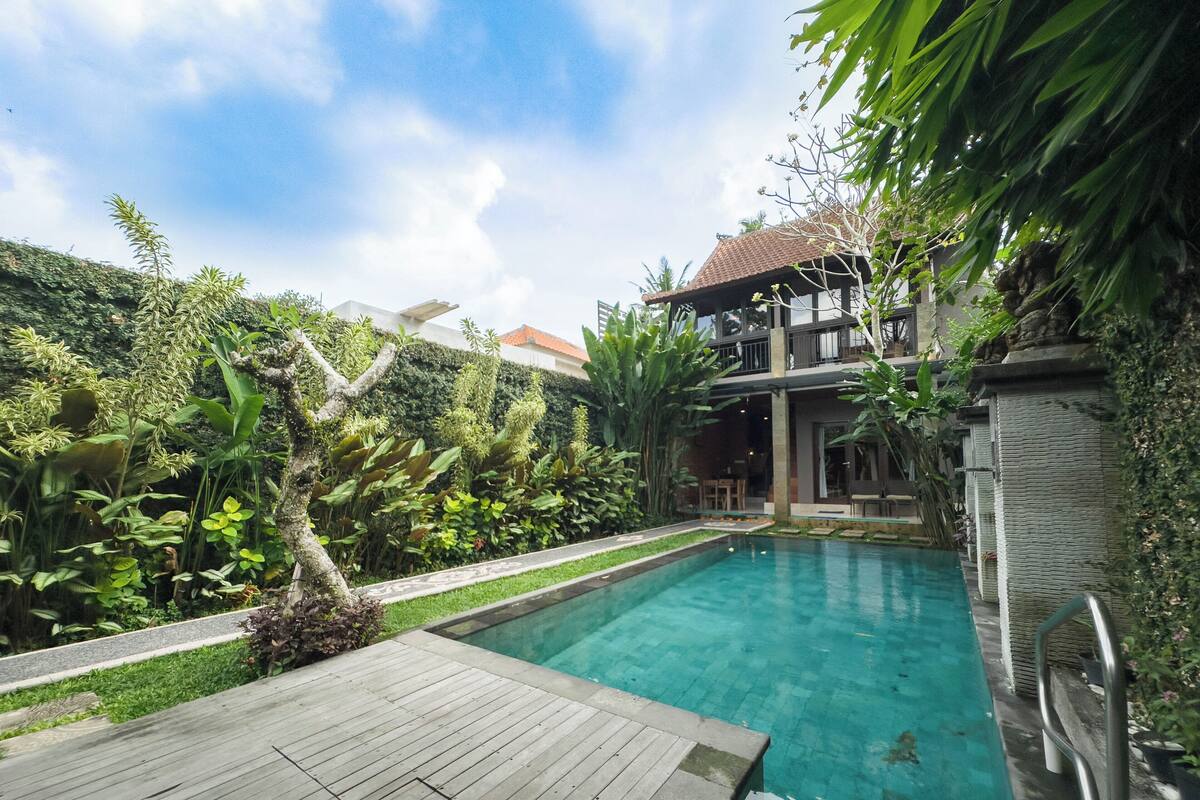 A swimming pool with a wooden deck, surrounded by lush greenery and a two-story building with a tiled roof.