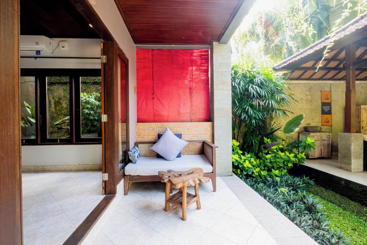 Un espacio al aire libre moderno con un banco acolchado y una pequeña mesa de madera, rodeado de abundante follaje y un muro rojo.