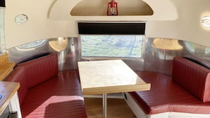 A compact RV interior with a dining area, two red cushioned benches, a small table, and a window with a view of greenery.