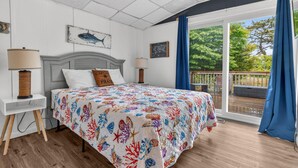 A bedroom with a bed, two bedside tables, a headboard, a window with curtains, and a view of an outdoor deck.