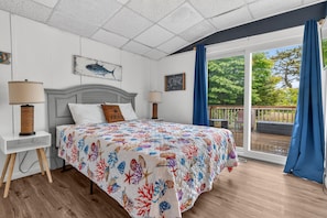 A bedroom with a bed, two bedside tables, a headboard, a window with curtains, and a view of an outdoor deck.