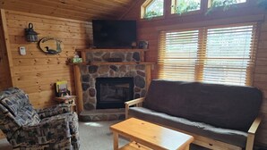 L’intérieur chaleureux d’un chalet avec une cheminée en pierre, un canapé, un fauteuil à motifs et un téléviseur fixé au mur.