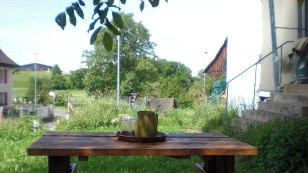 A wooden table with a green mug and a plate, set outdoors with plaid cushions. - Gite Rural, restaurant without alcohol/pork (Beurnevésin)