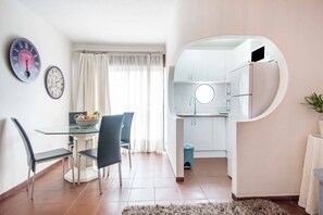 A compact kitchen with a refrigerator, a sink, and a round mirror. There is a dining area with a glass table and chairs, and a window with curtains. - Apartment Montechoro (Albufeira)