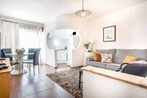 Een moderne woonkamer met een grijze bank, een eethoek met een glazen tafel en een keuken met witte kastjes.