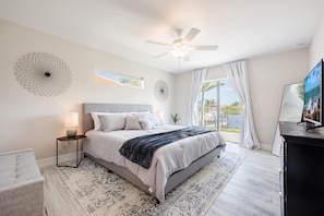A bedroom with a bed, a ceiling fan, a nightstand, a lamp, a TV, and a view of the outdoors. - The Charm of a sand castle, creative & artisitc, Waterfront - Villa Sand Castle - Roelens Vacations (Cape Coral)