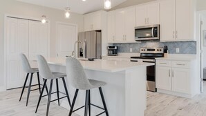 A modern kitchen with white cabinetry, a central island with bar stools, stainless steel appliances, and pendant lighting. - The Charm of a sand castle, creative & artisitc, Waterfront - Villa Sand Castle - Roelens Vacations (Cape Coral)