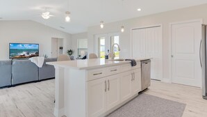 A modern kitchen with a central island, white cabinetry, and a built-in dishwasher. There is a grey sofa and a flat-screen TV in the living area. - The Charm of a sand castle, creative & artisitc, Waterfront - Villa Sand Castle - Roelens Vacations (Cape Coral)