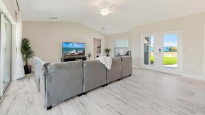 A spacious living room with a grey sofa, a flat-screen TV, and a ceiling fan. - The Charm of a sand castle, creative & artisitc, Waterfront - Villa Sand Castle - Roelens Vacations (Cape Coral)