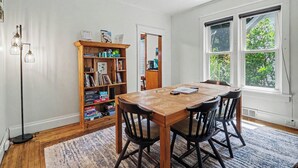 Uma área de refeições com uma mesa e cadeiras de madeira, uma estante de livros e uma janela com vista para o verde.