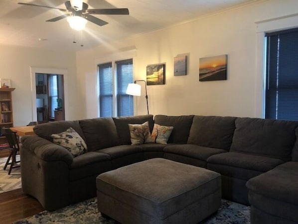 A living room with a sectional sofa, an ottoman, a ceiling fan, and artwork on the walls.