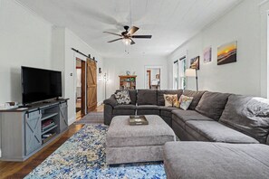 A living room with a sectional sofa, a flat-screen TV, a ceiling fan, and a large window.