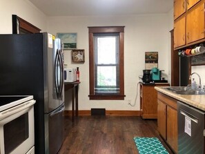 A compact kitchen with a refrigerator, microwave, stove, and sink. There is a window with blinds, wooden cabinets, and a small rug on the floor.