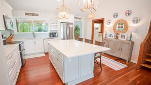 Una cocina con armarios blancos, una isla central, piso de madera, iluminación colgante y un comedor con mesa y sillas de madera.