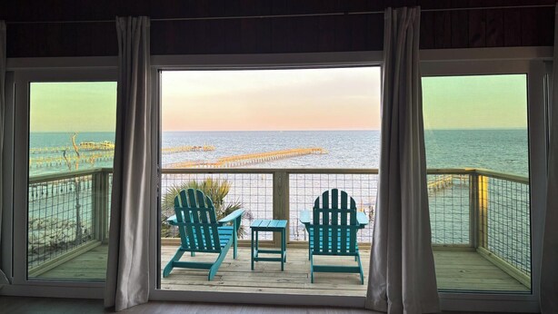 Una habitación con un ventanal grande que da a un muelle y al mar. Hay dos sillas Adirondack verdes y una mesita.