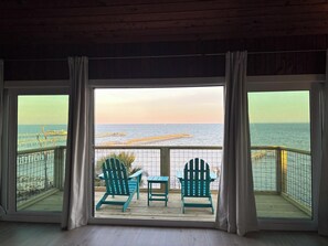 Quarto com uma grande janela com vista para um cais e o mar. Há duas cadeiras Adirondack verdes e uma pequena mesa.