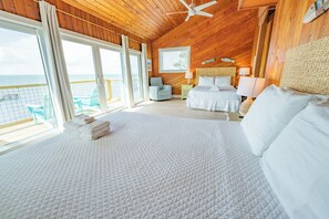 A bedroom with a large bed, two bedside tables, a ceiling fan, and a view of the ocean. - BAYFRONT - Old Florida Style Cottage w/ private dock! (St. George Island)
