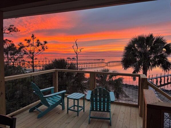 A wooden deck with two chairs and a small table, overlooking a sunset over water with a pier and palm trees. - BAYFRONT - Old Florida Style Cottage w/ private dock! (St. George Island)