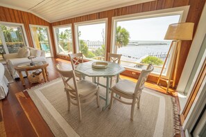 A room with a wooden floor, a round table with chairs, a sofa, and large windows offering a view of the sea and a pier. - BAYFRONT - Old Florida Style Cottage w/ private dock! (St. George Island)