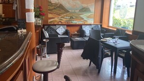 A restaurant with a bar area, seating with cushioned sofas and chairs, a large mural on the wall, and a window with a view of greenery.