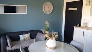 Ett rum med en mörkblå vägg, ett vitt runt bord, en svart soffa med mönstrade kuddar, ett vitt skåp och en väggklocka.