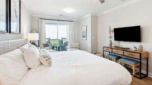 A hotel room with a large bed, a desk with drawers, a television, and a balcony with outdoor seating. - Beautiful Spacious Oceanfront Condo On Isle of Palms (Isle of Palms)