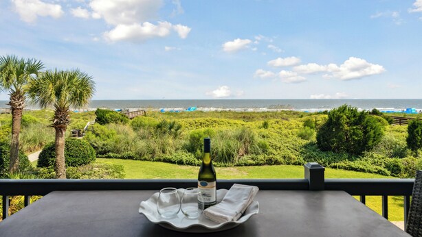 A balcony with a table, a bottle of wine, and glasses on a tray, overlooking a coastal landscape with greenery and the sea. - Beautiful Spacious Oceanfront Condo On Isle of Palms (Isle of Palms)