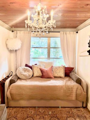 Ein Schlafzimmer mit einem Bett, einer Kristallleuchtre, einem Fenster mit Vorhängen und einem weißen, flauschigen Kissen an der Wand.