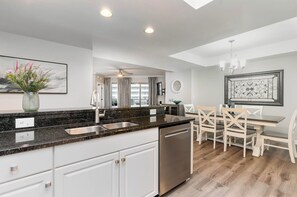 A modern kitchen with white cabinets, a granite countertop, and a dishwasher. There is a dining area with a table and chairs.