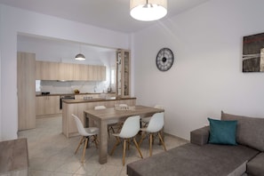 A modern kitchen with wooden cabinets, a dining area with a wooden table and white chairs, and a grey sofa with a teal pillow.