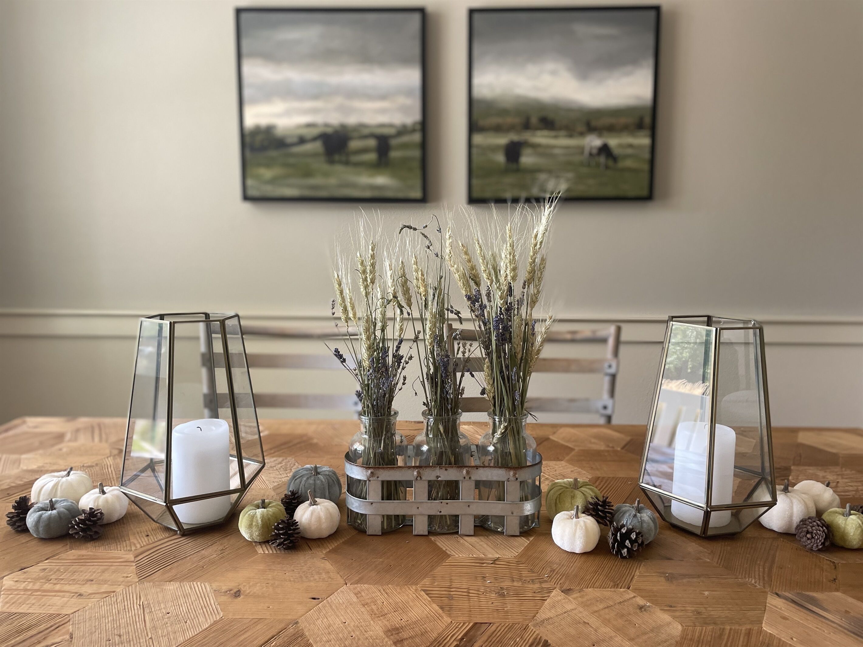 A dining table with two glass candle holders, a vase with dried flowers, and decorative pumpkins.