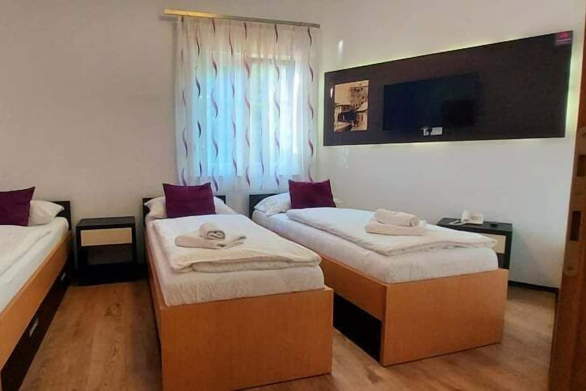 A hotel room with two single beds, a small table, a nightstand, a window with curtains, and a wall-mounted TV.