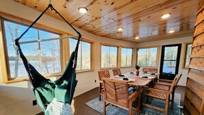 Ein Holzinterieur mit einem Hängekorbstuhl, einem Esstisch mit Stühlen und großen Fenstern mit Blick auf Bäume und ein Gewässer.