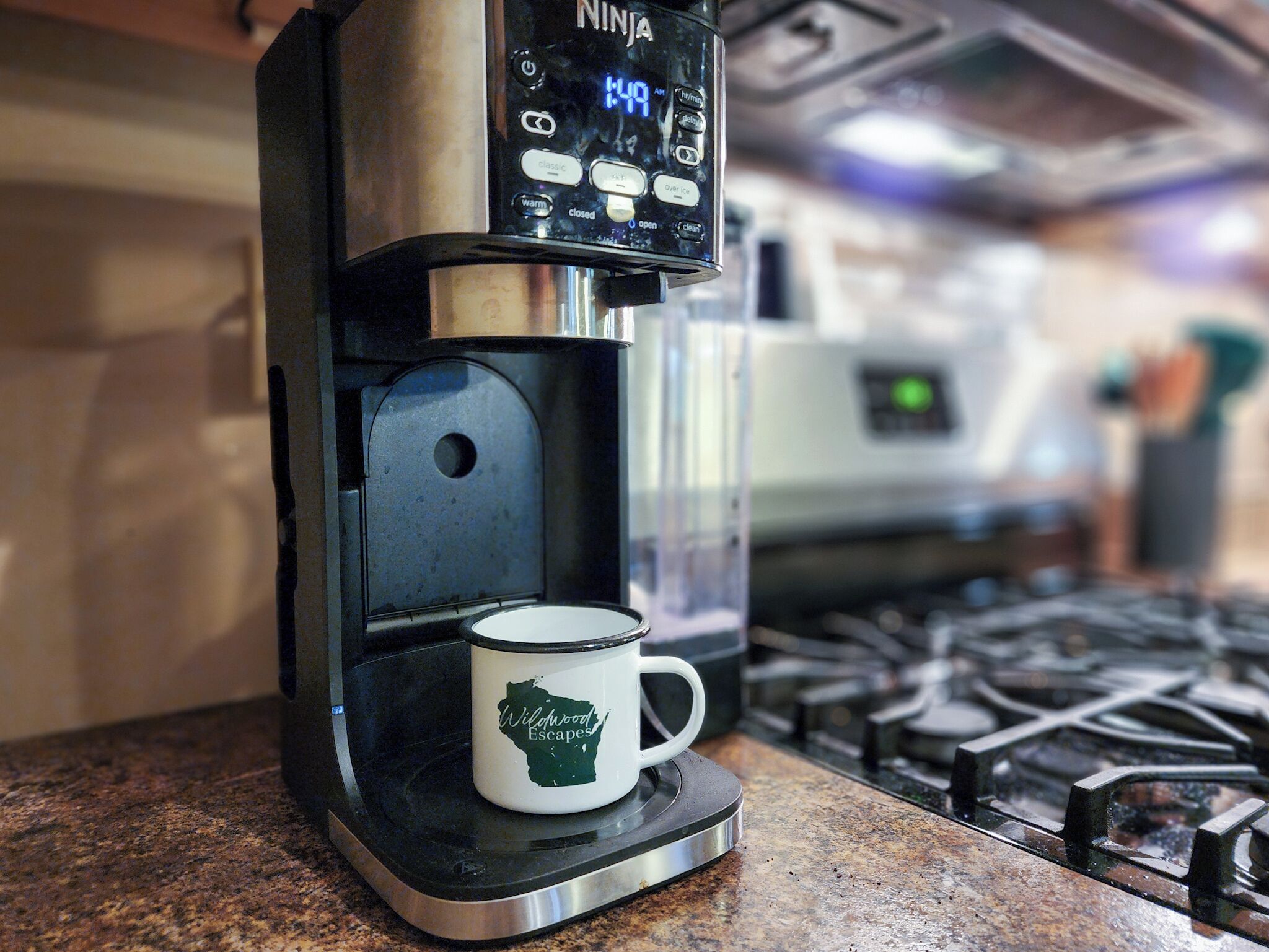 A Ninja coffee maker with a mug on its drip tray, a kitchen stove, and a microwave in the background.