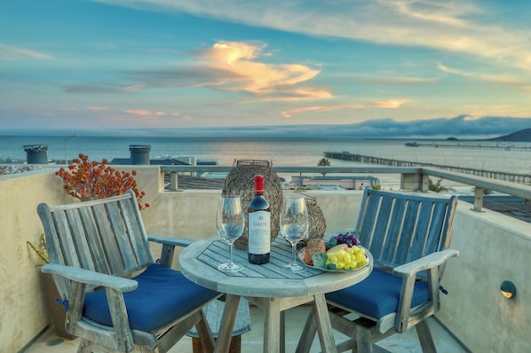 Una terraza con dos sillas de madera, una mesa redonda, una botella de vino y un cuenco de fruta.
