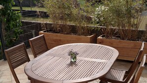 Een ronde houten tafel met stoelen op een betegeld terras, omgeven door potplanten en met uitzicht op een woonwijk.