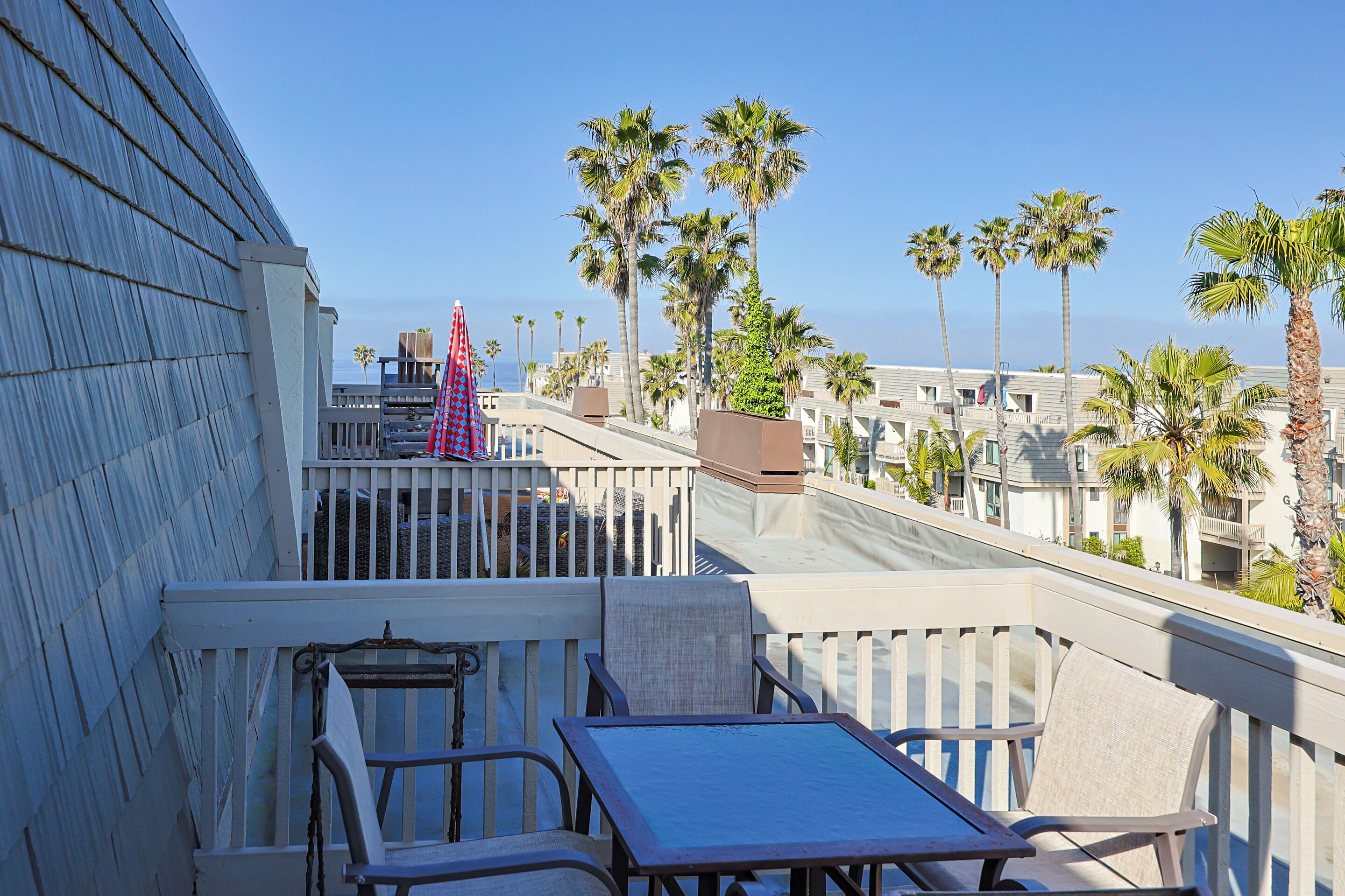Un balcon avec une table et des chaises, d’où l’on peut admirer une rangée de palmiers et un ciel bleu dégagé.