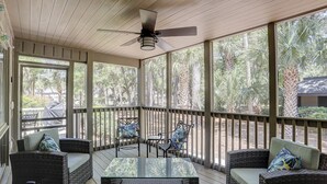 A covered outdoor seating area with wicker furniture and a glass-top table, overlooking a garden with palm trees.