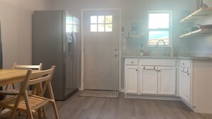 A compact kitchen with white cabinets, a stainless steel refrigerator, a dining table with chairs, and a window providing natural light. - Cheerful studio by LionsBridge, short 🚶‍♀️to downtown. (St. Augustine)