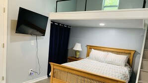 A single bedroom with a bed, a nightstand, a lamp, a TV mounted on the wall, and a window with curtains. - Cheerful studio by LionsBridge, short 🚶‍♀️to downtown. (St. Augustine)