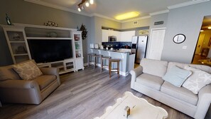 A living room with a TV, two sofas, a coffee table, and a kitchen area with a fridge, microwave, and bar stools.