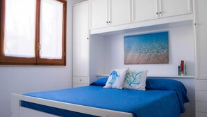 A bedroom with a bed, a window, white cabinets, and a painting of a beach scene. - Detached house in San Vito Lo Capo near the beach (San Vito Lo Capo)