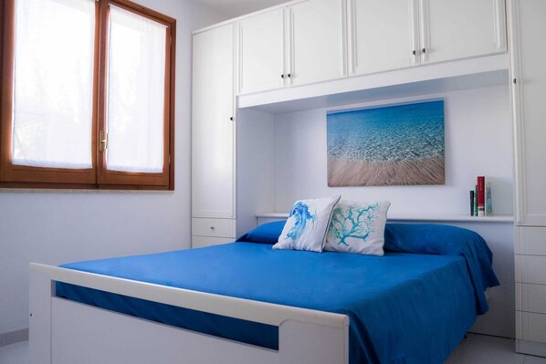 Un dormitorio con una cama, una ventana, armarios blancos y un cuadro de una playa.